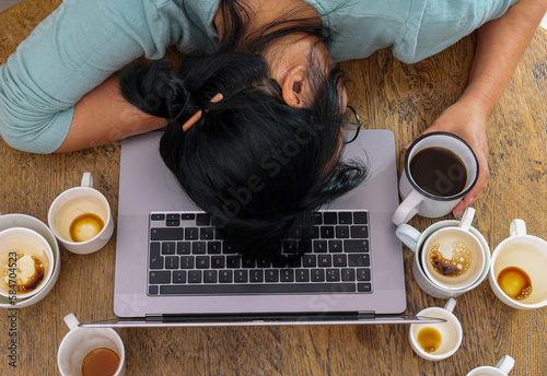 Obraz na plátne Exhausted female worker surrounded by coffee cups sleeping at workplace over laptop