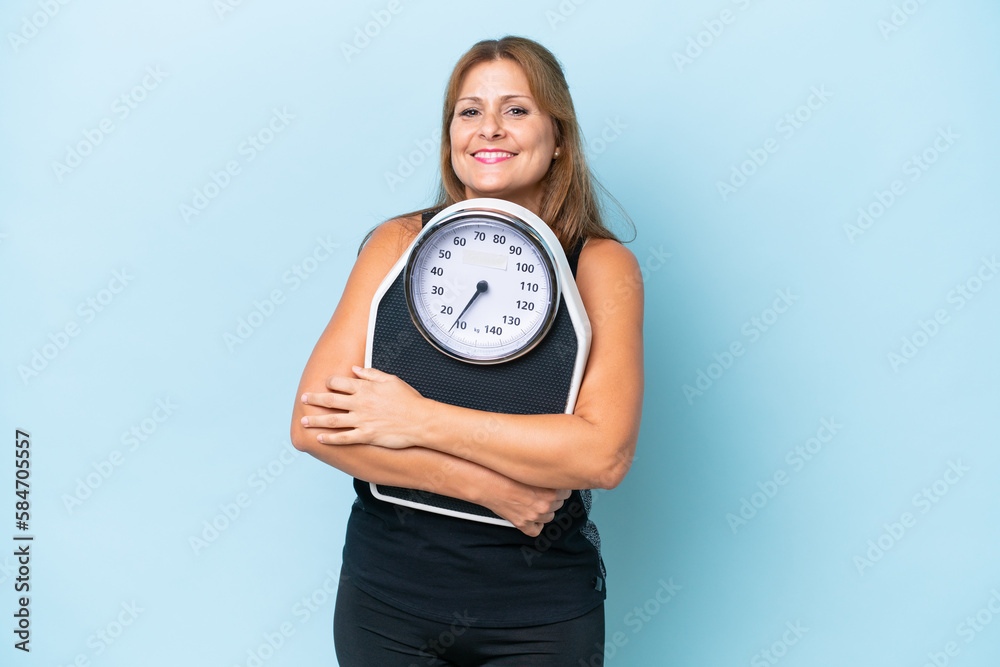 Middle-aged caucasian woman isolated on blue background with weighing machine Middle-aged caucasian woman isolated on blue background with weighing machine