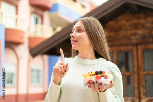 Young pretty blonde woman holding a bowl of fruit at outdoors intending to realizes the solution while lifting a finger up