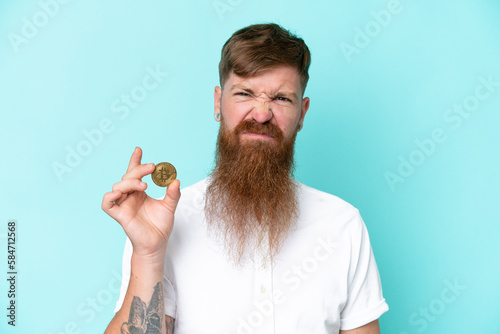 Redhead man with long beard holding a Bitcoin isolated on blue background with sad expression