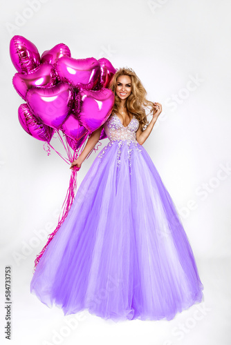 Beautiful woman in amazing long dress is holding pink balloons and is posing on white background. Elegant female model in gown dress. Elegance. Glamour.