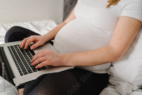 Closeup of pregnant woman writing notes and using a laptop while working on maternity leave in bedroom. Female have computer on knees  laps work as freelancer in bed at home. Remote work  overwork.