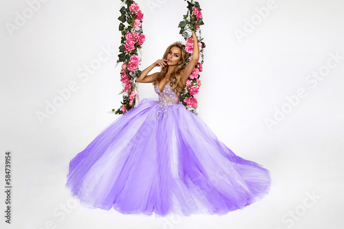 Beautiful natural woman in elegant long dress on a floral swing on white background. Glamour female model. Spring concept. Bridal fashion.
