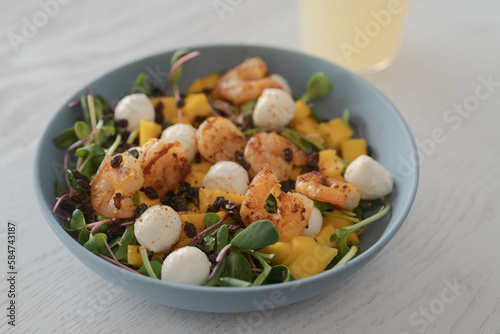 Salad with mango, mozzarella, shrimps and mixed microgreens in blue bowl on white oak table