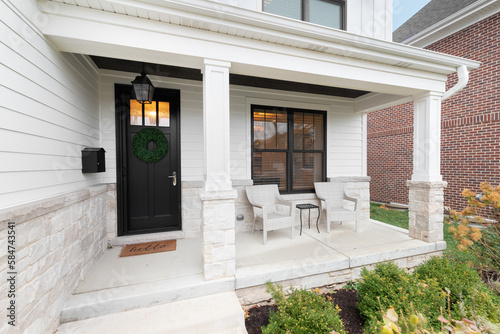 A new, white modern farmhouse with a dark wood door with windows, white pillars, a stone floor, and patio furniture.