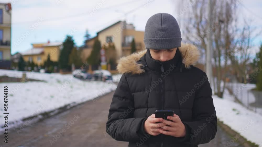 Young boy walking with mobile phone at winter day. Student holding smartphone. Checking email, writing typing sms, browsing social media. Outdoor, gadget using concept Young boy walking with mobile phone at winter day. Student holding smartphone. Checking email, writing typing sms, browsing social media. Outdoor, gadget using concept