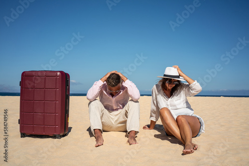 Lost couple of travelers sitting sad on the sandy beach with a big suitcase. Conceptual photo. photo