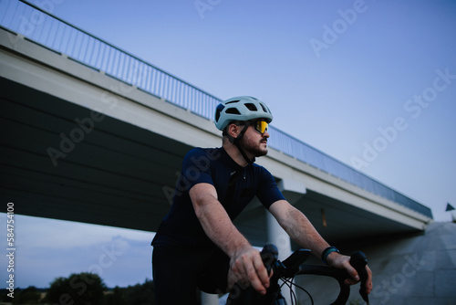 Close up of young man with strong body wearing cycling clothing and sneakers riding bike on paved road. Natural landscape and evening time.