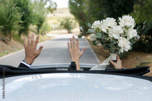 due neo sposi tirano in alto le mani ce il bouquet che esce dal tetto dell'auto in corsa photo