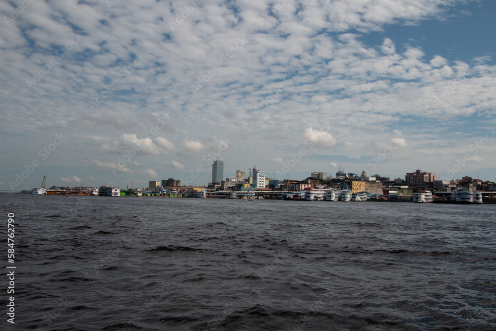 River view of a ferry refueling at a floating fuel station at the port of Manaus, with city skyscrapers visible in the background. Location: Manaus, Brazil