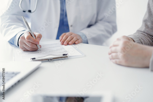 Doctor and patient sitting at the table in clinic. The focus is on female physician's hands filling up the medication history record form or checklist, close up. Medicine concept