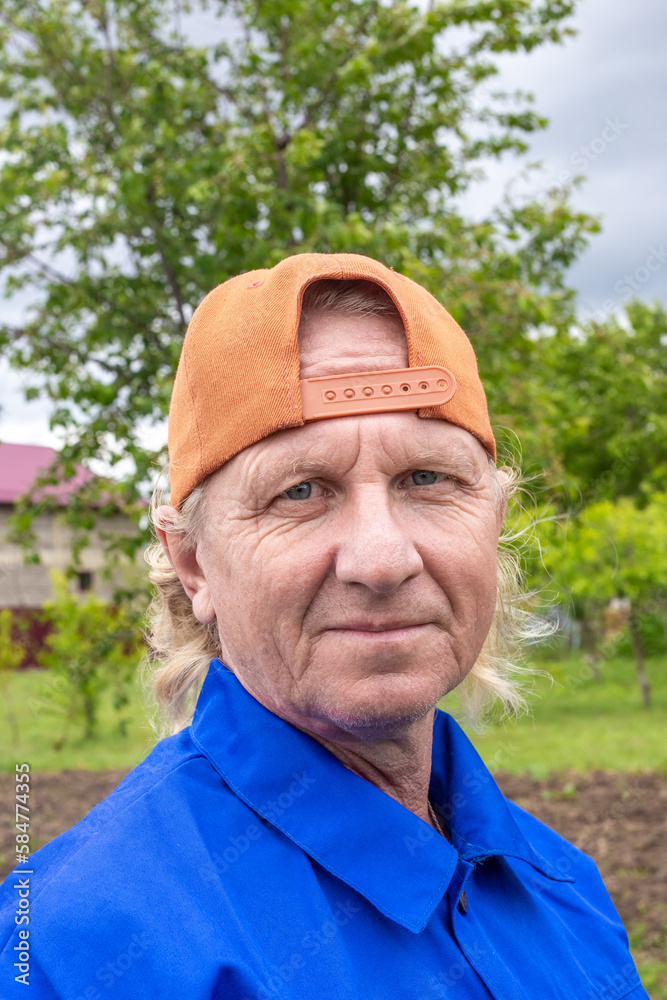 A working man in a blue jacket and cap looks at the camera in a garden in summer. Portrait of a Caucasian man.