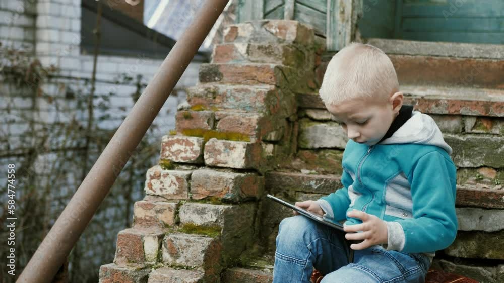 A handsome boy of 5 years old is watching a video or playing a mobile game on a tablet while sitting on the steps of a dilapidated, shabby, dilapidated building. A handsome boy of 5 years old is watching a video or playing a mobile game on a tablet while sitting on the steps of a dilapidated, shabby, dilapidated building.