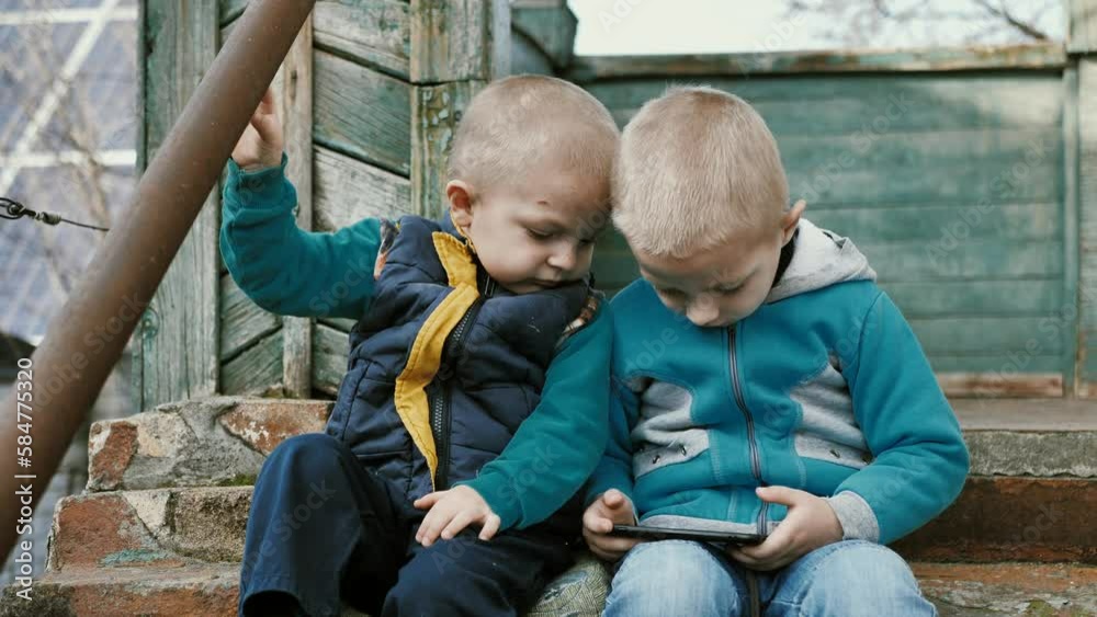 Portrait of children looking at mobile phone. The brothers watch videos or play video games on their mobile phones while sitting on the steps of a dilapidated building. Children's gadget