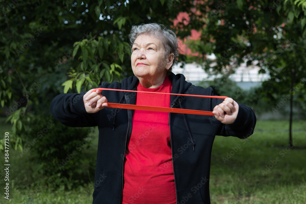 Smiling elderly woman is doing exercises with fitness elastic band outdoors in the yard. Active life of pensioner. Adaptation of pensioners in modern world. Prevention of brain diseases. Mental health