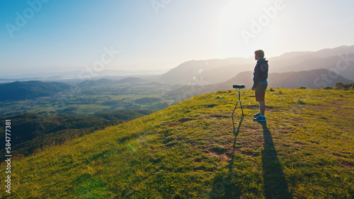 Photographer on the hill. Young male photographer takes pictures with camera and telephoto lens set on tripod on the green hill in the mountains at sunrise