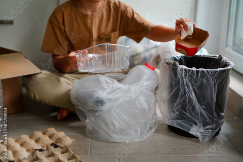 a boy of eight years old in beige clothes, sits at home in the kitchen and sorts garbage into different bags for recycling, the child separates plastic and kraton, bottles and bags