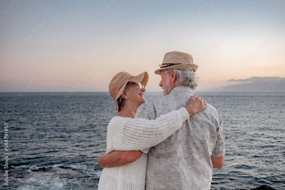 Back view of romantic senior couple or pensioners embraced at the sea at sunset light expressing love and tenderness - old couple outdoors enjoying vacations together