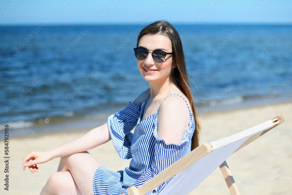 Happy young brunette woman relaxing on a wooden deck chair at the ocean beach while looking at camera, smiling, and wearing fashion sunglasses. The enjoying vacation concept