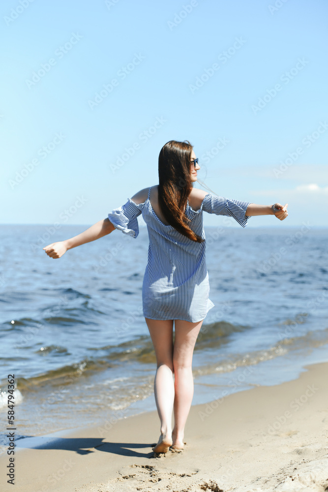 Happy smiling woman in free bliss on ocean beach standing with open hands. Portrait of a brunette female model in summer dress enjoying nature during travel holidays vacation outdoors, back view