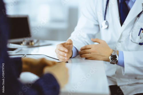 A doctor is talking to his patient  while sitting together at the desk in the cabinet in a hospital. Physician using clipboard for filling up medication history records. Perfect medical service