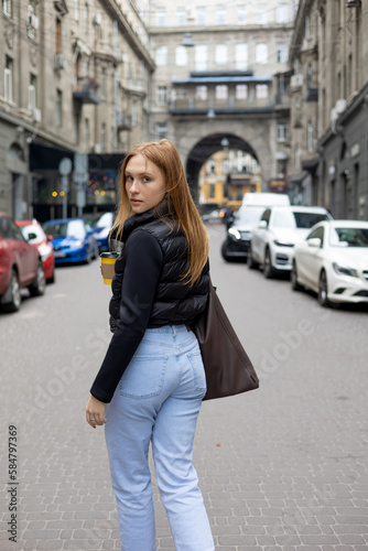 stylish young female in jeans and coat strolling along street near modern building and drinking coffee © Mykola