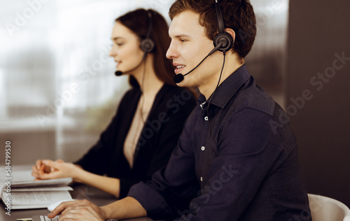 Two young people in headsets are talking to the clients, while sitting at the desk in a modern office. Focus on man in a blue shirt. Call center operators at work