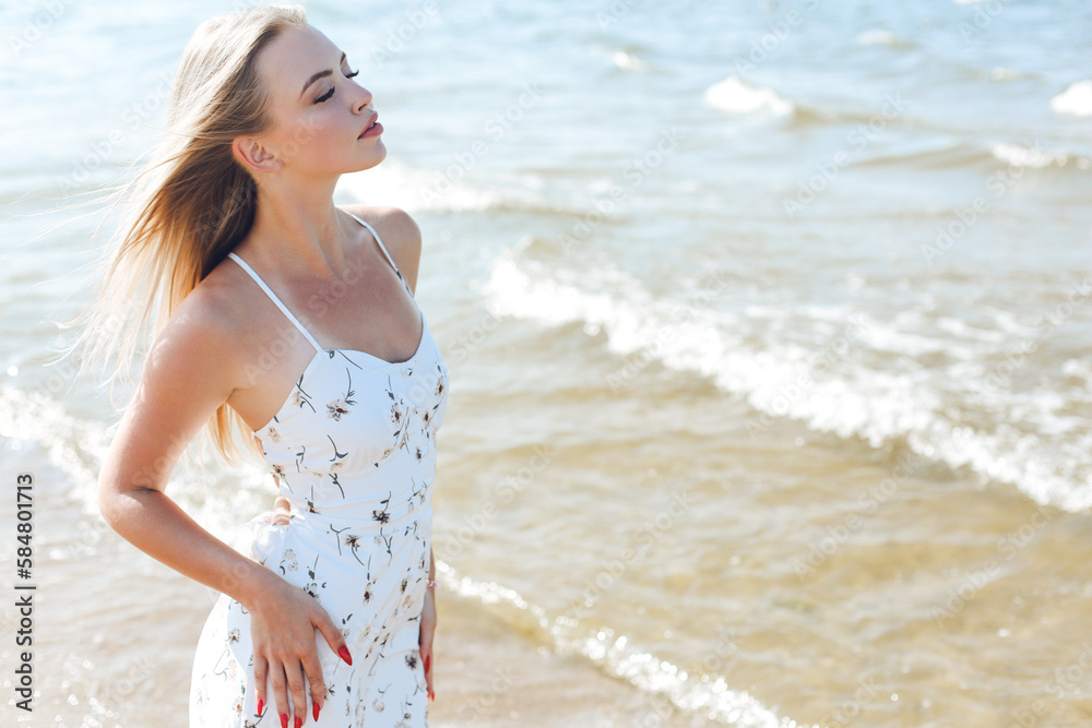 Happy blonde woman in free happiness bliss on ocean beach standing straight. Portrait of a female model in white summer dress enjoying nature during travel holidays vacation outdoors