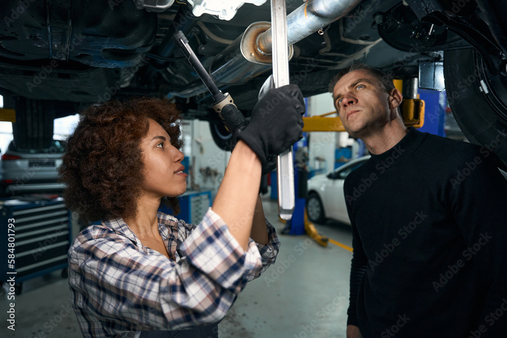 Young woman auto mechanic and client inspect car from below Young woman auto mechanic and client inspect car from below
