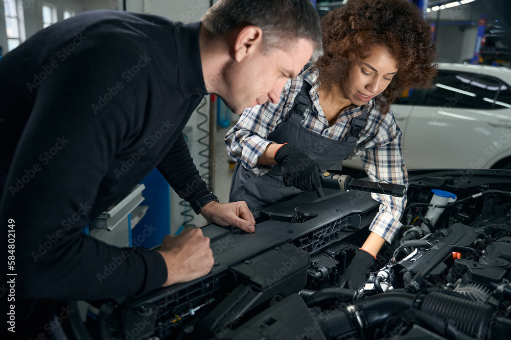 Man in casual clothes and a woman in work overalls look under the hood of a car Man in casual clothes and a woman in work overalls look under the hood of a car