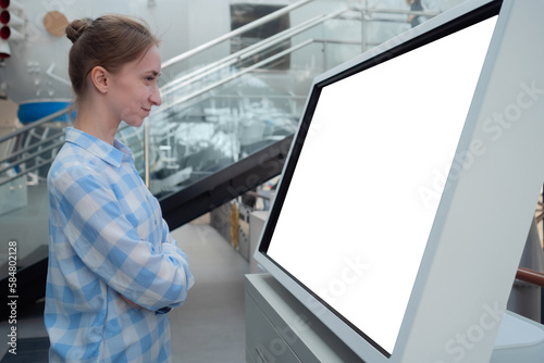 Side view: woman in blue plaid shirt looking at blank interactive horizontal white display of electronic kiosk at futuristic exhibition or museum. White screen, mock up, template, technology concept.