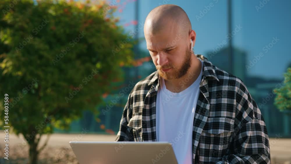 Handsome Man Sitting on the Bench Outside, Working on Laptop, Using Earphones. Businessman Is Remote Working with Wireless Portable Computer, Typing, Online Chatting, Browsing, and Programming Apps. Handsome Man Sitting on the Bench Outside, Working on Laptop, Using Earphones. Businessman Is Remote Working with Wireless Portable Computer, Typing, Online Chatting, Browsing, and Programming Apps.