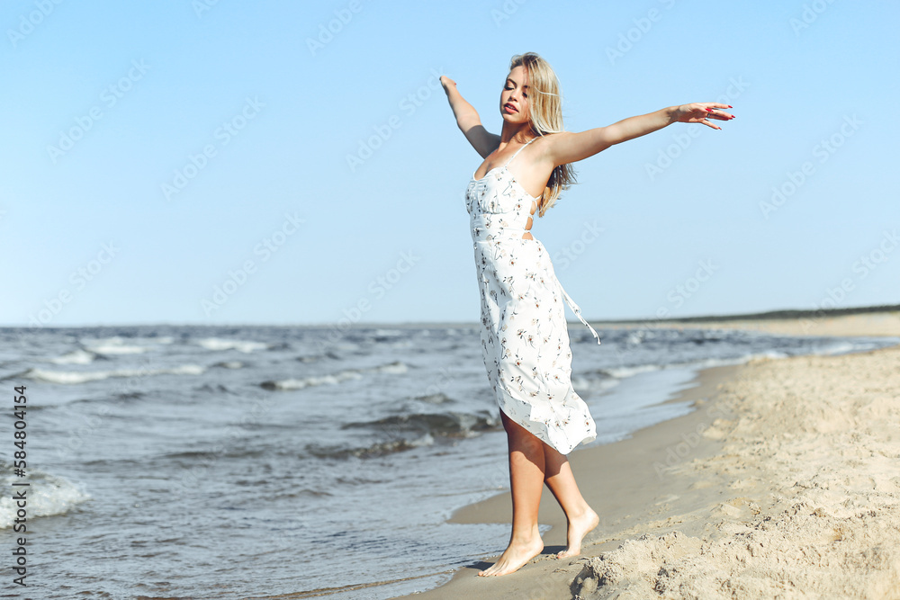Happy blonde beautiful woman on the ocean beach standing in a white summer dress, open arms Happy blonde beautiful woman on the ocean beach standing in a white summer dress, open arms