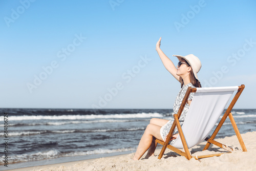 Happy brunette woman sitting on a wooden deck chair at the ocean beach while waving and greeting somebody with her hand © rogerphoto