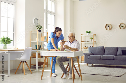 Demented senior man spends time in retirement home. Friendly nurse helping old man who is playing games and doing puzzles while sitting at desk in cozy retirement home interior. Dementia care concept