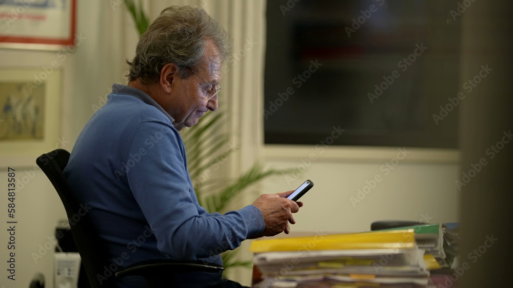 Fototapeta premium Candid mature man looking at smartphone device at home office. One older perosn in 70s working using cellphone reading message