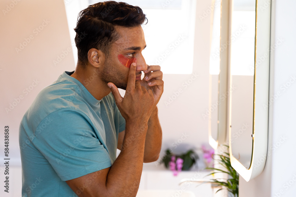 Biracial man looking in mirror and applying under eye mask in bathroom Biracial man looking in mirror and applying under eye mask in bathroom