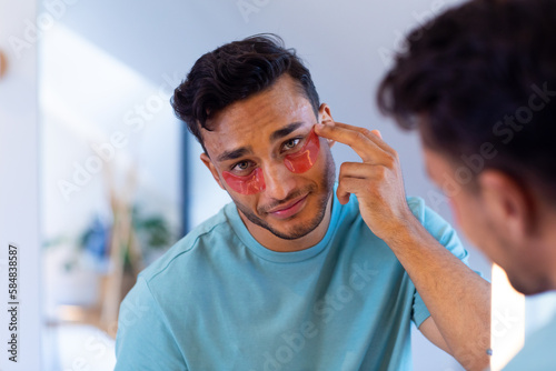 Biracial man looking in mirror and applying under eye masks in bathroom