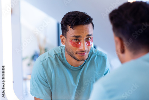 Smiling biracial man with under eye masks looking in mirror in bathroom