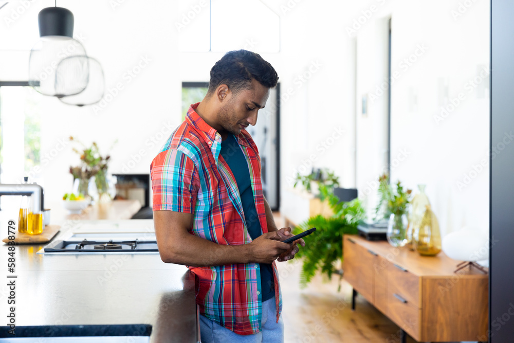 Happy biracial man leaning on countertop and using smartphone in kitchen Happy biracial man leaning on countertop and using smartphone in kitchen