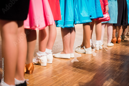 Young girls ballroom dancers standing in a row before beginners competition. Colorful dresses and different shoes on wooden floor. Kids ballroom dancing.