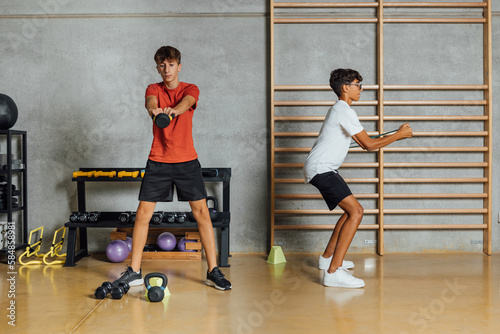 Full length of Boys Teenagers exercising circuit routine, training with dumbbells at gym with concrete wall. Workout, lifting weight, jumping. Horizontal.