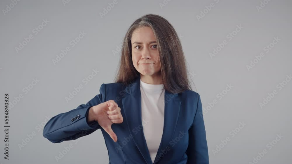 Attractive middle aged caucasian brunette business woman wearing blue jacket shows thumbs down dislike sign standing on grey background, beautiful female office manager looking at camera, studio shot Attractive middle aged caucasian brunette business woman wearing blue jacket shows thumbs down dislike sign standing on grey background, beautiful female office manager looking at camera, studio shot