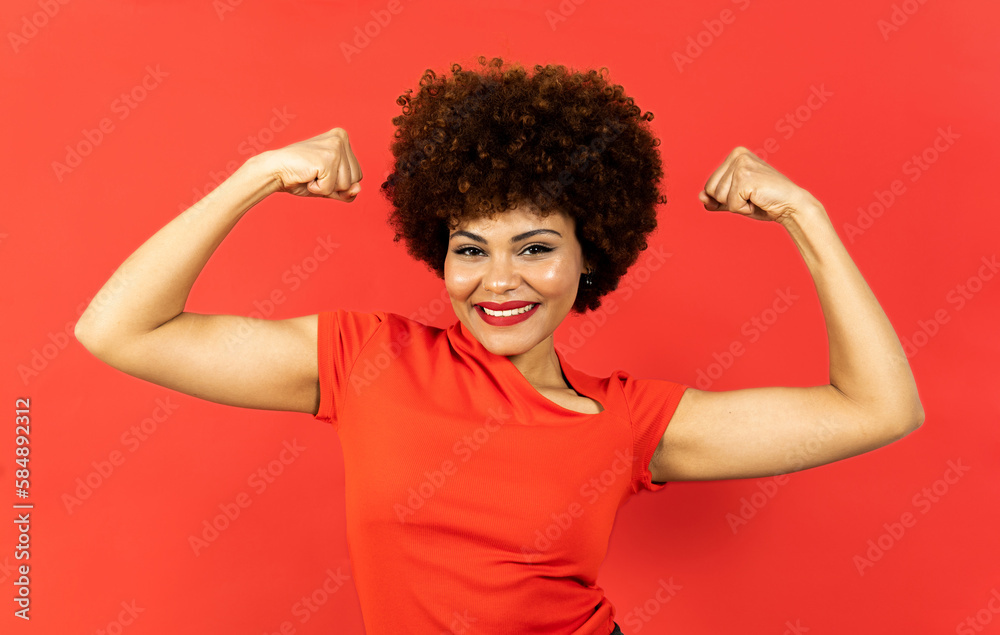 A dark-skinned woman with afro hair poses on a red background. The girl shows the two biceps with expression of encouragement, winner or motivation. Concept of empowered woman, overcoming. A dark-skinned woman with afro hair poses on a red background. The girl shows the two biceps with expression of encouragement, winner or motivation. Concept of empowered woman, overcoming.