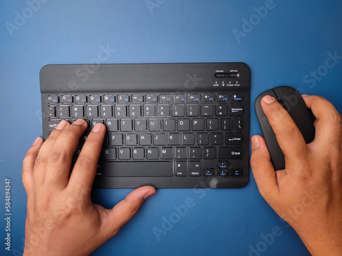 Top view someone hand typing wireless keyboard with holding mouse on blue background.