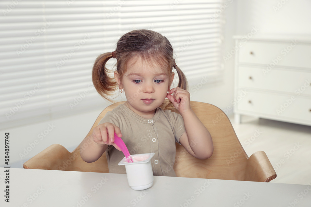 Cute little child eating tasty yogurt from plastic cup with spoon at white table indoors Cute little child eating tasty yogurt from plastic cup with spoon at white table indoors