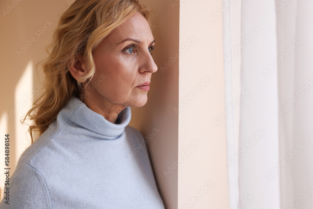 Upset middle aged woman near window at home. Loneliness concept Upset middle aged woman near window at home. Loneliness concept