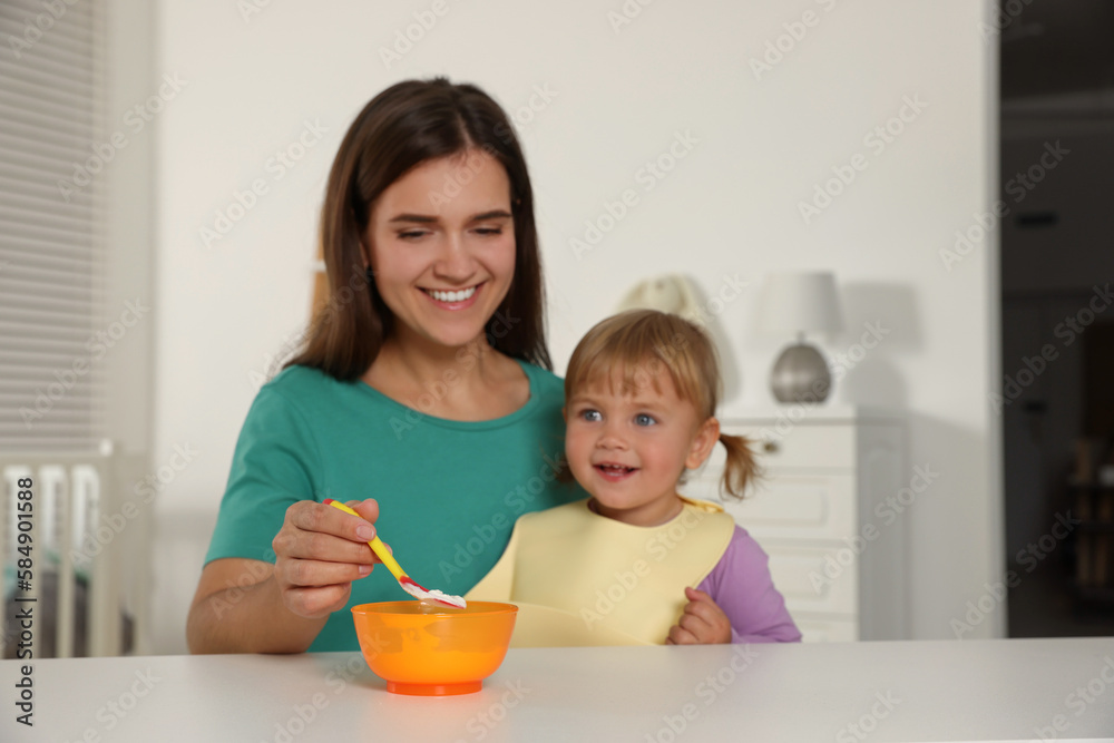 Mother feeding her cute little child with yogurt at white table in room Mother feeding her cute little child with yogurt at white table in room