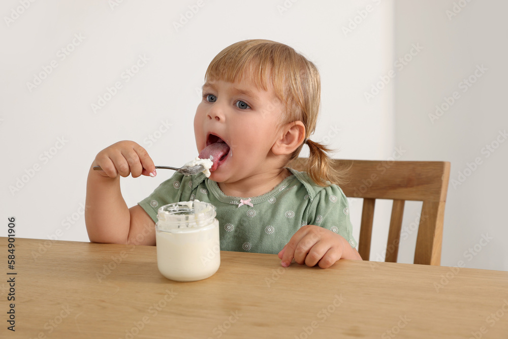 Cute little child eating tasty yogurt with spoon at wooden table indoors Cute little child eating tasty yogurt with spoon at wooden table indoors