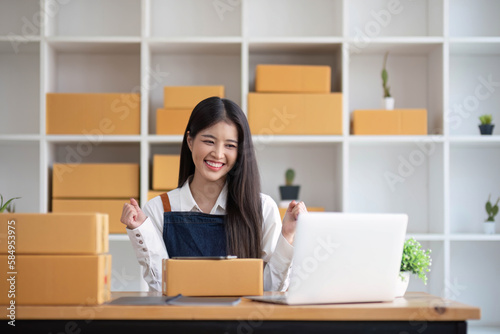 A portrait of a young Asian woman, e-commerce employee sitting in the office full of packages in the background write note of orders and a calculator, for SME business ecommerce and delivery business © wichayada